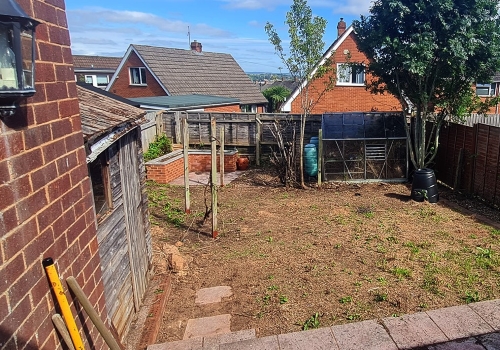 Garden cleared of brambles and vegetation, revealing a once covered greenhouse, shed and raised bed around a lawn and patio.